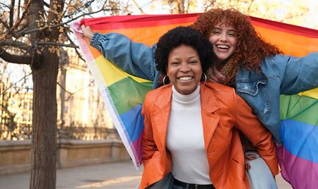 A person is giving a piggyback ride to another person holding a rainbow flag in an urban park setting.